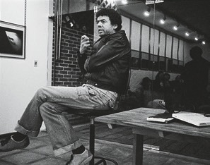 Descrição da imagem: Fotografia em preto e branco de George Love sentado de lado em uma cadeira alta, usando jaqueta de couro, calça jeans e sapatos. Ele gesticula com as mãos e olha para frente. Ao fundo, há uma parede de tijolos e o reflexo em uma parede de vidro de uma fileira de pessoas sentadas a sua frente. À direita, uma mesa com alguns objetos em cima, incluindo papéis e livros. Fim da descrição da imagem.