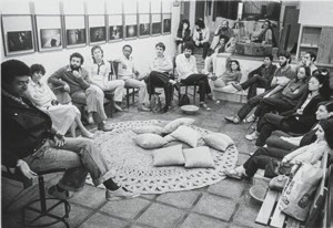 Descrição da imagem: Fotografia em preto e branco e horizontal. Um grupo de aproximadamente vinte pessoas está reunido em uma sala, sentadas em cadeiras e sofás dispostos em um círculo. No centro do círculo há um tapete com almofadas. A maioria das pessoas está olhando para George Love, posicionado nesta roda na parte inferiro esquerda da imagem. As paredes da sala são decoradas com várias fotografias alinhadas em fileiras horizontais. Fim da descrição da imagem.