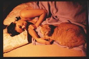 Descrição da imagem: Fotografia colorida e horizontal de Rosi deitada de lado em uma cama, sob um cobertor lilás. Ela acaricia um gato laranja deitado ao seu lado, que está sobre a coberta. Fim da descrição da imagem.