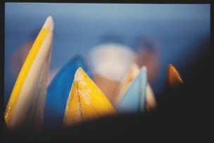 Descrição da imagem: Fotografia colorida e horizontal. A imagem mostra várias pontas de pranchas de surfe coloridas em primeiro plano, com cores como amarelo, azul e branco. As pranchas estão agrupadas e em uma posição vertical, apontando para cima. O fundo da imagem está desfocado, mostrando silhuetas indistintas de pessoas e um céu azul, a praia e o mar ao fundo. A desfocagem do fundo destaca as pranchas de surfe em primeiro plano. Fim da descrição da imagem.