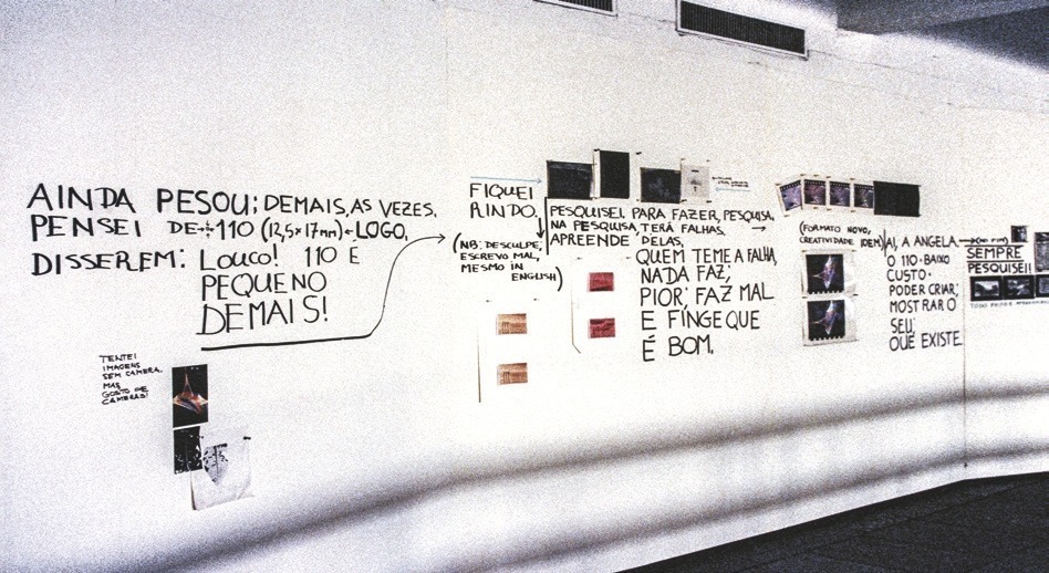"Descrição da imagem: Fotografia colorida de uma parede branca coberta por anotações escritas à mão em tinta preta e colagens de fotos. As frases escritas incluem reflexões, notas e pensamentos sobre pesquisa e processos criativos. Algumas das frases dizem: ""AINDA PENSOU; DEMAIS, AS VEZES. PENSEI DE: ± 110 (12,5 x 17 cm) e – LOGO, DISSEREM: Louco! 110 é PEQUENO DEMAIS!"" ""Tentei imagens sem camera, mas gosto de cameras."" ""FIQUEI RINDO - (NB: de sculpe, escrevo mal, mesmo in English) ""PESQUISEI PARA FAZER, PESQUISA, NA PESQUISA, TERÁ FALHAS, APREENDE DELAS."" ""QUEM TEME A FALHA, NADA FAZ, PIOR; FAZ MAL E FINGE QUE É BOM."" ""(Formato novo, Creatividade IDEM)"" Entre as anotações, há várias pequenas fotografias e imagens coladas na parede, criando uma composição interligando imagens e escritas por setas. Fim da descrição da imagem."