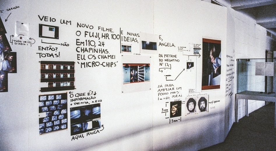 Descrição da imagem: Fotografia colorida de uma parede branca coberta com diversas fotografias, negativos e anotações manuscritas em preto. No topo da parede, há o texto: "VEIO UM NOVO FILME. O FUJI, HR 100". Abaixo desse texto, há várias imagens pequenas e tiras de negativos presos na parede. Há anotações que explicam o conteúdo das imagens e negativos, incluindo frases como "EU OS CHAMEI 'MICRO-CHIPS'" e "E NOVAS IDEIAS". À direita, há um retrato colorido de uma pessoa e mais notas, incluindo "E, ANGELA" e diagramas que destacam partes específicas de fotos. A parede está cheia de setas e linhas que conectam as imagens às anotações. Fim da descrição da imagem.