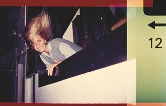 Descrição da imagem: Fotografia colorida de Ângela, uma pessoa com cabelos loiros esvoaçantes inclinada, apoiando o tronco em uma janela, sorrindo. A pessoa veste uma camisa branca com listras verticais e um colete azul acizentado. Ela está em em um ambiente externo, com prédios ao fundo. À direita da imagem, há uma borda colorida com a numeração "12" e uma seta apontando para a esquerda. Fim da descrição da imagem.