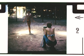 Descrição da imagem: Fotografia colorida de uma criança Yanomami de pé ao centro da imagem, com o torso nu, e em um ambiente ao ar livre com chão de terra batida. À direita, em primeiro plano, Cláudia Andujar está agachada de costas, segurando uma câmera fotográfica em um tripé e tirando uma foto da criança. Ela está com as costas a mostra e usa calça jeans. Ao fundo, pontos de luz solar pentram entre as árvores. A foto está inserida em um retângulo com bordas brancas e moldura preta e marcas de numeração na lateral direita, com um 9 e uma seta apontando para a esquerda. Fim da descrição da imagem.