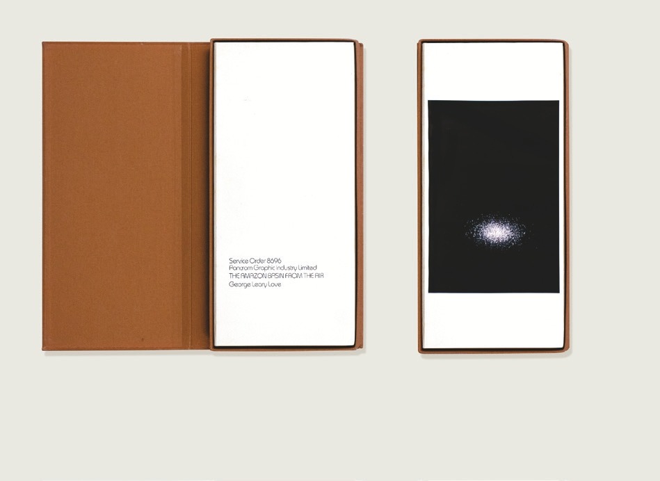 Descrição da imagem: A imagem mostra o estojo do Livro "Service Order 8696" e duas de suas imagens. À esquerda, o estojo está aberto com a capa marrom a mostra. À direita, apenas a imagem interna é visível. Elas estão dispostas lado a lado sobre uma superfície branca. O livro à esquerda tem a página em branco com o texto "Service Order 8606 Panorama Graphic Industry, Limited THE AMAZON RIVER SEEN FROM THE AIR George Leary Love" na parte inferior da página. No livro à direita, uma fotografia das águas de um rio com o reflexo do sol em forma circular iluminando pontualmente a parte inferior da imagem, em um alto contrate de do escuro das águas com o claro do sol. Fim da descrição da imagem.