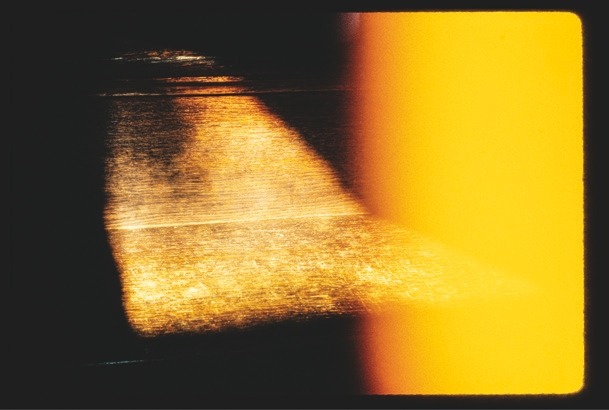 Descrição da imagem: Fotografia colorida abstrata com predominância de tons escuros e amarelos. A imagem apresenta um contraste forte entre áreas escuras à esquerda e uma faixa vertical larga em amarelo brilhante à direita. No centro, há uma zona de transição onde a luz amarela parece iluminar uma superfície texturizada, criando um efeito de reflexo e brilhos dourados. Fim da descrição da imagem.