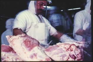 Descrição da imagem: Fotografia colorida de um trabalhador em uma linha de processamento de carne. O homem está usando um uniforme branco, incluindo touca, máscara e avental, e está manuseando grandes pedaços de carne crua sobre uma superfície de trabalho. À esquerda e à direita, outros trabalhadores também estão presentes, embora parcialmente fora de foco. O ambiente parece ser um frigorífico ou uma fábrica de processamento de alimentos, com iluminação artificial e atmosfera industrial. Fim da descrição da imagem.