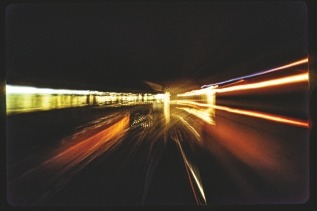 Descrição da imagem: Fotografia noturna abstrata de dois trens se cruazando em uma linha. Traços de luzes coloridas em movimento, formando linhas que se estendem a partir do centro da imagem em direção às bordas, sugere o efeito de velocidade e distorção. As luzes são predominantemente amarelas e vermelhas. Fim da descrição da imagem.