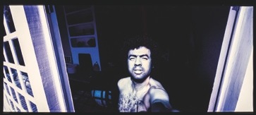 Descrição da imagem: Fotografia colorida de George Love tirando uma selfie embaixo de uma janela ou porta. Ele está sem camisa, iluminado por uma forte luz natural e seu rosto está parcialmente sombreado. Ao fundo, o ambiente interno é pouco iluminado, com alguns objetos e paredes parcialmente visíveis. Fim da descrição da imagem.