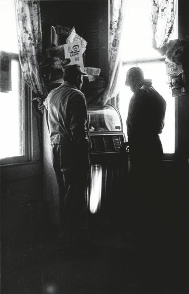Descrição da imagem: Imagem em preto e branco e vertical. A imagem mostra dois homens de pé, vistos de costas e ligeiramente de perfil, em frente a uma jukebox em um ambiente interno. Os homens estão próximos um do outro, olhando para o aparelho. A luz entra por duas janelas ao fundo, iluminando parcialmente a cena e criando silhuetas marcadas em grande contraste. Cortinas com padrões florais e prateleiras com objetos diversos são visíveis na área superior. Fim da descrição da imagem.