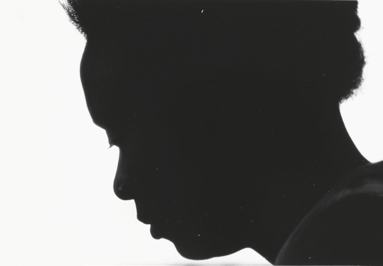 Descrição da imagem: Fotografia em preto e branco, mostrando a silhueta do perfil de uma pessoa negra, de cabelo curto e crespo. A imagem tem a silhueta da pessoa destacando-se contra um fundo claro, quase branco. A posição da cabeça inclinada para baixo, com a linha do queixo suavemente curvada, e sugere um momento de introspecção. Fim da descrição da imagem.