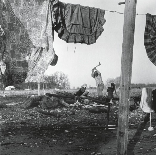 Descrição da imagem: Fotografia em preto e branco de uma cena rural. Em primeiro plano, roupas estão penduradas em um varal. Ao fundo, uma pessoa está cortando lenha com um machado, cercada por troncos de árvores já cortados e espalhados pelo chão. Fim da descrição da imagem.