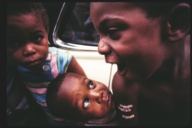 Descrição da imagem: Uma fotografia em cores mostrando o rosto de três crianças negras com um carro ao fundo. À esquerda, uma criança observa para frente, com um olhar curioso. No centro, outra criança olha atentamente para cima, observando a terceira criança. Esta, à direita, está gritando ou falando com intensidade, de perfil, virada para a esquerda. A imagem é dinâmica e transmite a ideia de interação entre as crianças. Fim da descrição da imagem.