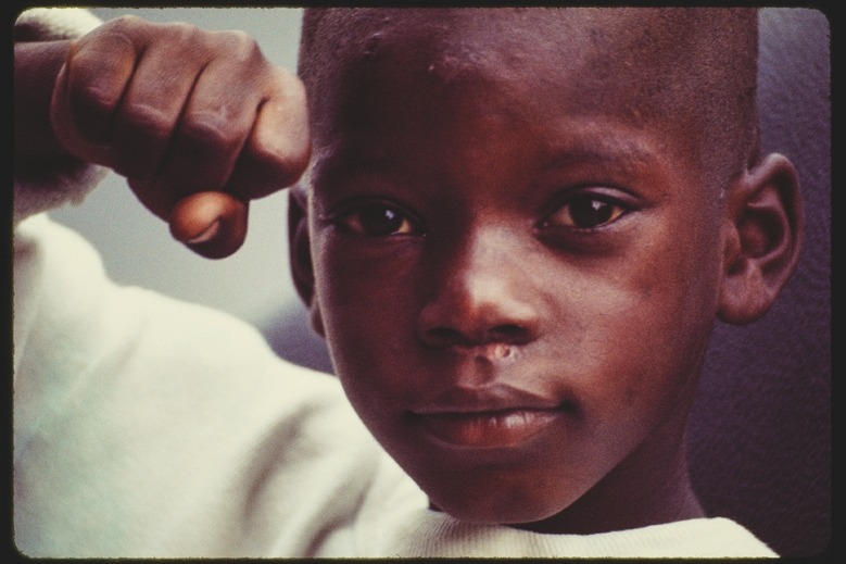 Descrição da imagem: Uma fotografia colorida do rosto de um menino negro que encara a câmera com uma expressão confiante. Ele está com o braço levantado e dobrado para frente, com a mão fechada em um punho. A iluminação destaca seu rosto e olhos. Ele usa uma blusa branca comprida, tem a cabeça raspada e a marca de uma cicatriz logo abaixo do nariz. A imagem foca em sua expressão e no gesto do punho, com o fundo desfocado. Fim da descrição da imagem.