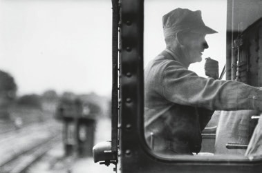 Descrição da imagem: A fotografia em preto e branco captura um maquinista de perfil dentro de uma cabine de trem. Ele está vestindo um uniforme com boné, e parece estar segurando um rádio ou algum dispositivo de comunicação próximo à boca, como se estivesse falando. A janela da cabine emoldura a cena. Ao fundo, desfocado, é possível ver trilhos de trem que se estendem até o horizonte, com outro vagão distante. A imagem parece enfatizar a concentração e o trabalho do maquinista no controle da locomotiva. Fim da descrição da imagem.