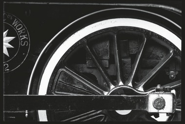 Descrição da imagem: Fotografia em preto e branco destaca uma roda de trem. O foco da imagem está na grande roda metálica, com raios robustos que suportam seu aro externo e a biela paralela ao chão. Parte da estrutura mecânica do trem também é visível na imagem, como barras e componentes de conexão. No canto esquerdo da imagem, há parte de um logotipo com um símbolo de estrela dentro de um círculo, vista parcialmente, e um texto ao redor, parcialmente legível, com a palavra "Works" circundando a estrela. Fim da descrição da imagem.
