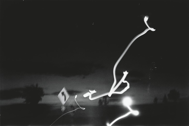 Descrição da imagem: Esta fotografia em preto e branco de um cenário noturno. Linhas de luz curvadas atravessam a imagem, resultado de uma longa exposição fotográfica que capturou o movimento de luzes em um ambiente escuro, técnica conhecida como "lightpainting". No fundo borrado, uma paisagem de estrada, com poucas árvores. À esrqueda da imagem há uma placa de trânsito " vire à esquerda" , indicando uma curva à frente. Fim da descrição da imagem.