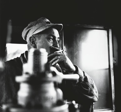 Descrição da imagem: Fotografia em preto e branco. Um homem de perfil usa um boné e fuma um cigarro. Ele está em um ambiente interno, com objetos desfocados em primeiro plano. A iluminação vem de uma janela ao fundo, criando um contraste entre luz e sombra no rosto do homem. A imagem transmite uma atmosfera contemplativa. Fim da descrição da imagem.