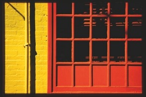 Descrição da imagem: Imagem colorida na horizontal de uma parede de tijolos amarelos e uma grande janela vermelha. À esquerda, sobre a parede, desce um cano também amarelo. À direita, a janela tem formato retangular, toda preenchida em quadrados. A parte inferior é fechada e a parte superior é vazada com vidros. Fim da descrição da imagem.