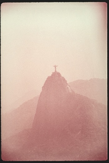 Descrição da imagem: Fotografia em tons de sépia. No centro, a silhueta do Cristo Redentor vista ao longe no topo do Corcovado, no Rio de Janeiro. A estátua e o morro estão envoltos por uma aparente neblina. O céu e o fundo se fundem em tons suaves. A imagem tem bordas escurecidas, sugerindo uma fotografia antiga. Fim da descrição da imagem.