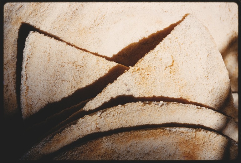 Descrição da imagem: A imagem apresenta uma série de fatias de um pão redondo, ao estilo pita, cortado em diversas fatias triangulares, sobrepostas um a um em camadas. A textura é áspera e arenosa, com uma coloração amarelada suave e que sugere uma superfície seca. As sombras entre as fatias criam um contraste marcante, destacando as formas triangulares. Fim da descrição da imagem.