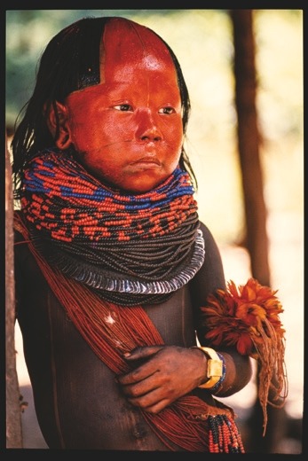 Descrição da imagem: Fotografia colorida vetical. Retrato de uma criança do povo Xikrin, da Amazônia. Seu rosto está pintado com tinta vermelha brilhante, cobrindo toda a parte superior da cabeça, que está raspada até o topo, até o início do peito. A criança veste várias camadas de colares de contas coloridas ao redor do pescoço, formando um padrão intrincado em cores azul, vermelho e preto. Ela também usa um bracelete dourado no pulso e um ramo de penas vermelhas no braço. O corpo, pintado de preto, é cruzado por um grande feixe de colares de contas vermelhas. Ela está apoiada em um poste de madeira. Fim da descrição da imagem.