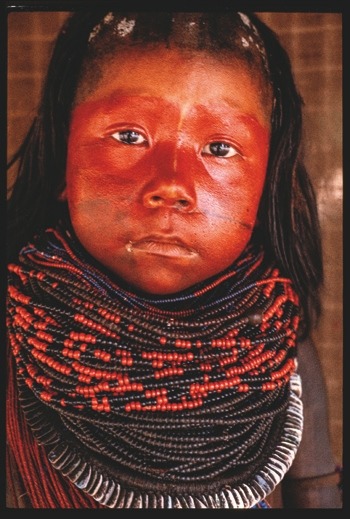 Descrição da imagem: Fotografia colorida vetical. Retrato de uma criança do povo Xikrin, da Amazônia, com o rosto pintado de vermelho. Ela usa uma série de colares de contas em tons de preto, vermelho e azul que cobrem boa parte do peito. O fundo da imagem é desfocado e de cor marrom. O cabelo da criança é escuro, raspado até o topo da cabeça e cai em volta do rosto pela parte de trás. Fim da descrição da imagem.