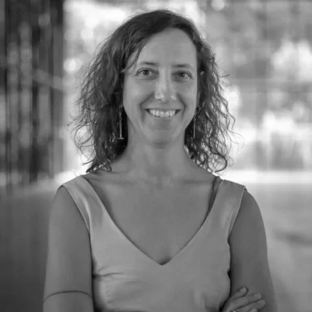 Início da descrição de imagem: Retrato frontal em preto e branco de uma mulher de pele clara, com cabelos castanhos ondulados e na altura dos ombros. Ela olha diretamente para a câmera com um sorriso largo e amigável. Veste uma blusa ou vestido de alças largas com decote em "V" e está com os braços cruzados na altura do peito. Em seus dedos, nota-se um anel largo de metal. O fundo da imagem está desfocado, sugerindo um ambiente arquitetônico amplo com colunas e luz natural, possivelmente as dependências de um museu. Fim da descrição de imagem.