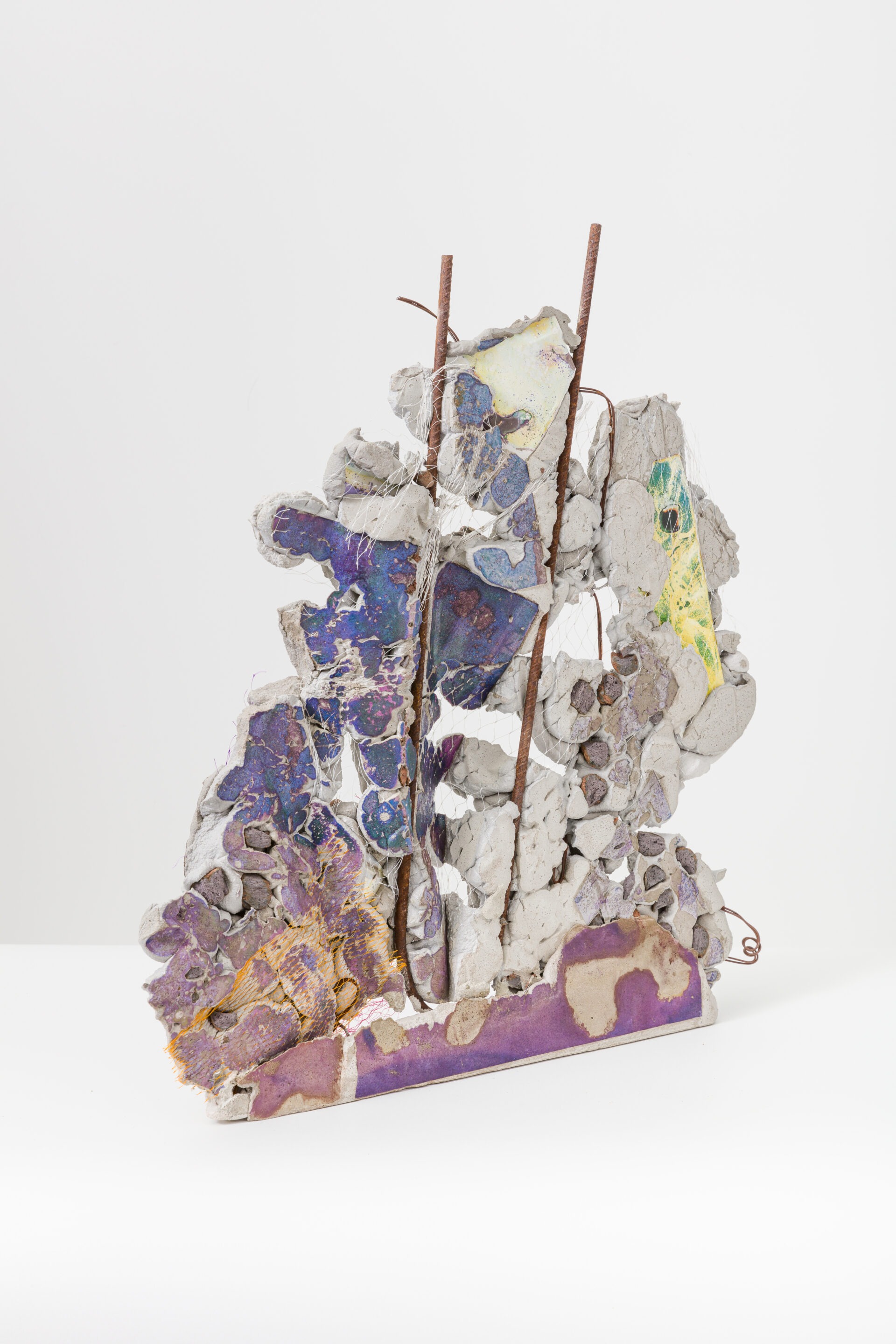 Descrição da imagem: Fotografia de uma escultura abstrata, vertical e assimétrica construída com pedaços de vários formatos e tamanhos de material cimentício em tons de roxo, branco e amarelo, entremeados por vergalhões enferrujados. Fim da descrição da imagem.