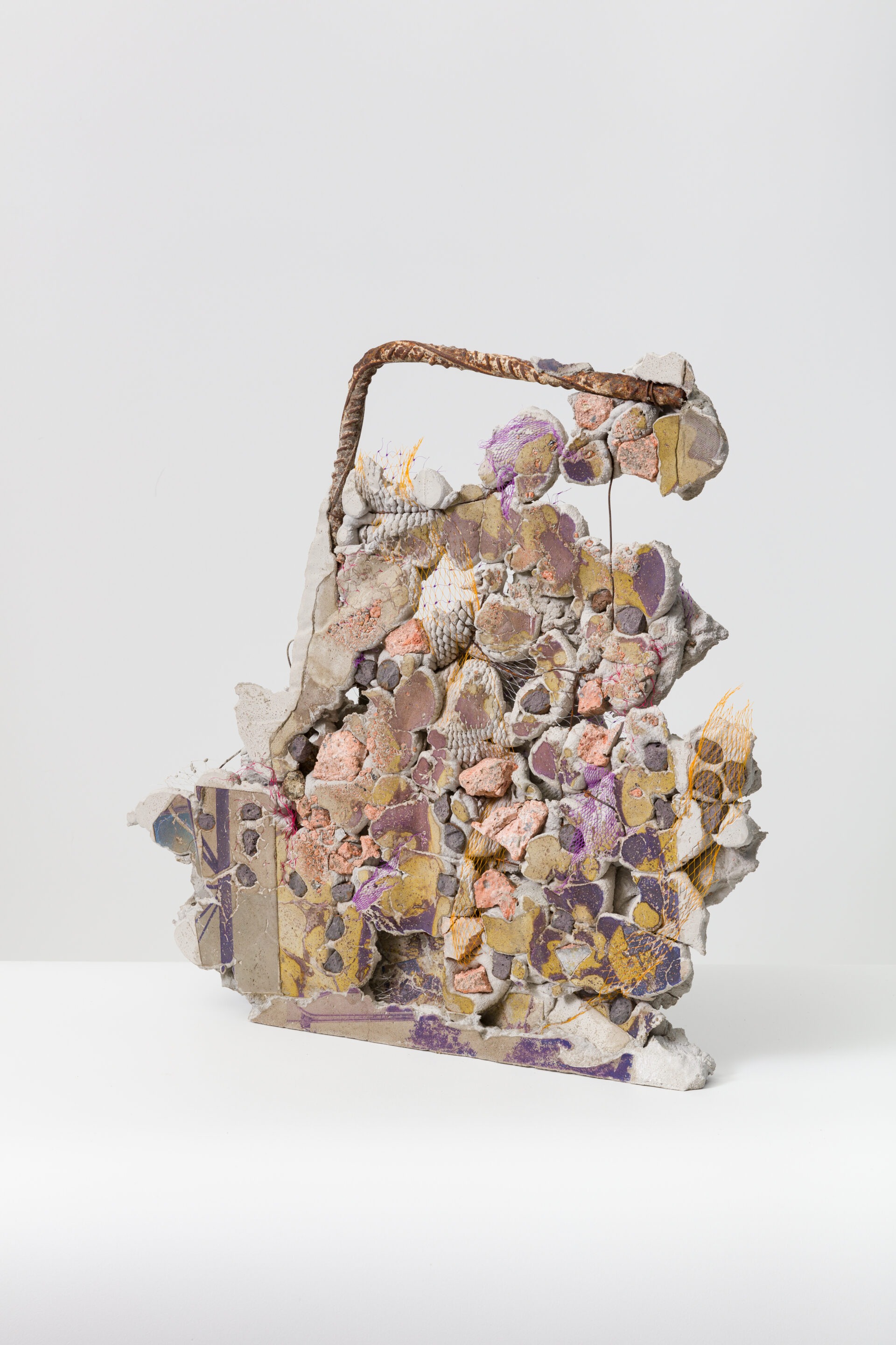 Descrição da imagem: Fotografia de uma escultura abstrata, vertical e assimétrica construída com pedaços de vários formatos e tamanhos de material cimentício em tons de laranja, rosa e roxo. No topo, à esquerda, um vergalhão enferrujado com a ponta inclinada para a direita. Fim da descrição da imagem.