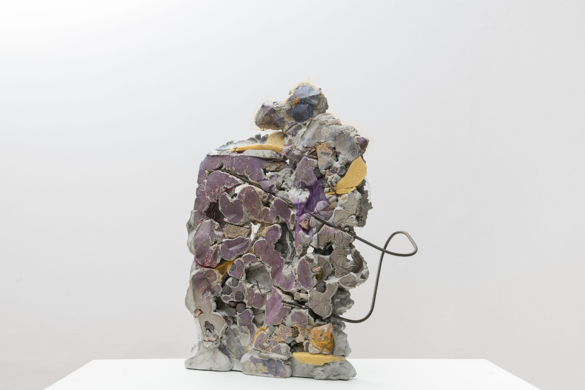 Descrição da imagem: Fotografia de uma escultura abstrata, vertical e assimétrica construída com pedaços de vários formatos e tamanhos de material cimentício em tons de cinza, laranja e roxo.Da estrutura se projeta pela direita um fino vergalhão enferrujado e retorcido. Fim da descrição da imagem.