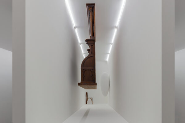 Descrição de imagem: A foto apresenta uma instalação arquitetônica. Os objetos presentes na imagem são de madeira, com exceção de um objeto oval branco. A obra mostra três objetos dentro de um espaço branco e estreito, entre eles: uma cadeira de escritório de imigração, simbolizando a transição e a espera. Além disso, um cadeiral de igreja católica do século 19, que evoca o papel da religião na exploração colonial. Também está presente a escultura 