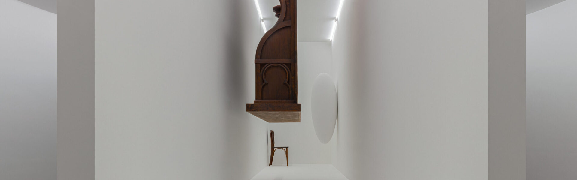 Descrição de imagem: A foto apresenta uma instalação artística. Os objetos presentes na imagem são de madeira, com exceção de um objeto oval branco. A obra mostra três objetos dentro de um espaço branco e estreito, entre eles: uma cadeira de escritório de imigração, simbolizando a transição e a espera. Além disso, um cadeiral de igreja católica do século 19, que evoca o papel da religião na exploração colonial. Também está presente a escultura "Cradle" ["Berço"] de Kiswanson, que aponta para o que está ausente. Esses elementos, flutuando fora de alcance, representam os limiares institucionais (igreja, escritório de imigração) que estruturam e tentam nomear vidas em transição como imigrações, exílios e colonizações. A obra desenvolve uma linguagem escultórica que se conecta à experiência do desterro do artista e à realidade global de diásporas e conflitos. Fim da descrição.