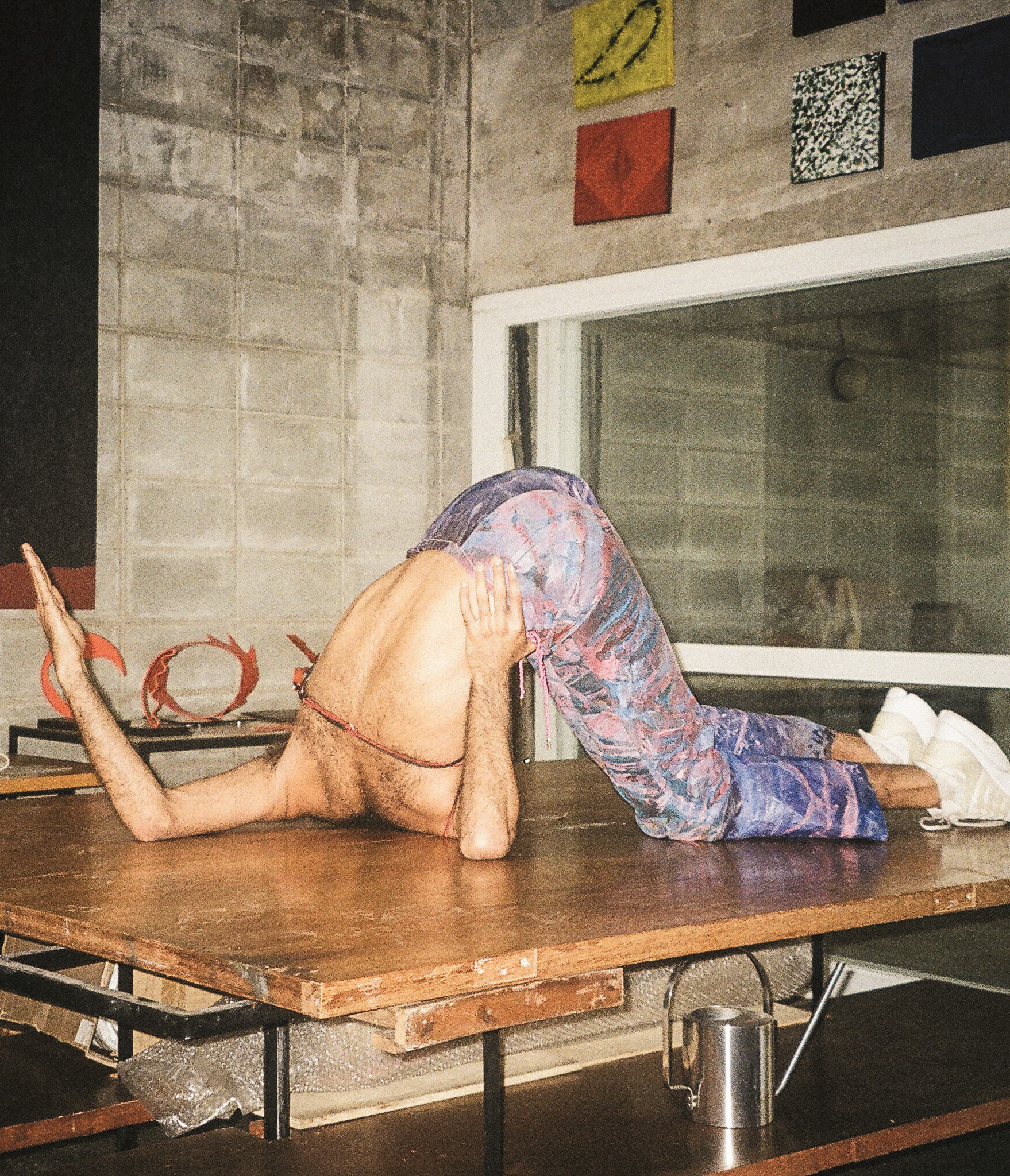 Descrição da imagem: Fotografia vertical de uma cena da Performance “Rumor”. Sobre uma mesa de madeira, uma pessoa negra de costas com o pescoço, os braços e as pernas apoiados nela, prestes a dar uma cambalhota. Está com o dorso desnudo e usa calça estampada de azul e roxo. Ao fundo, em uma parede de concreto, pequenos quadros coloridos dispostos de forma assimétrica. Fim da descrição da imagem.