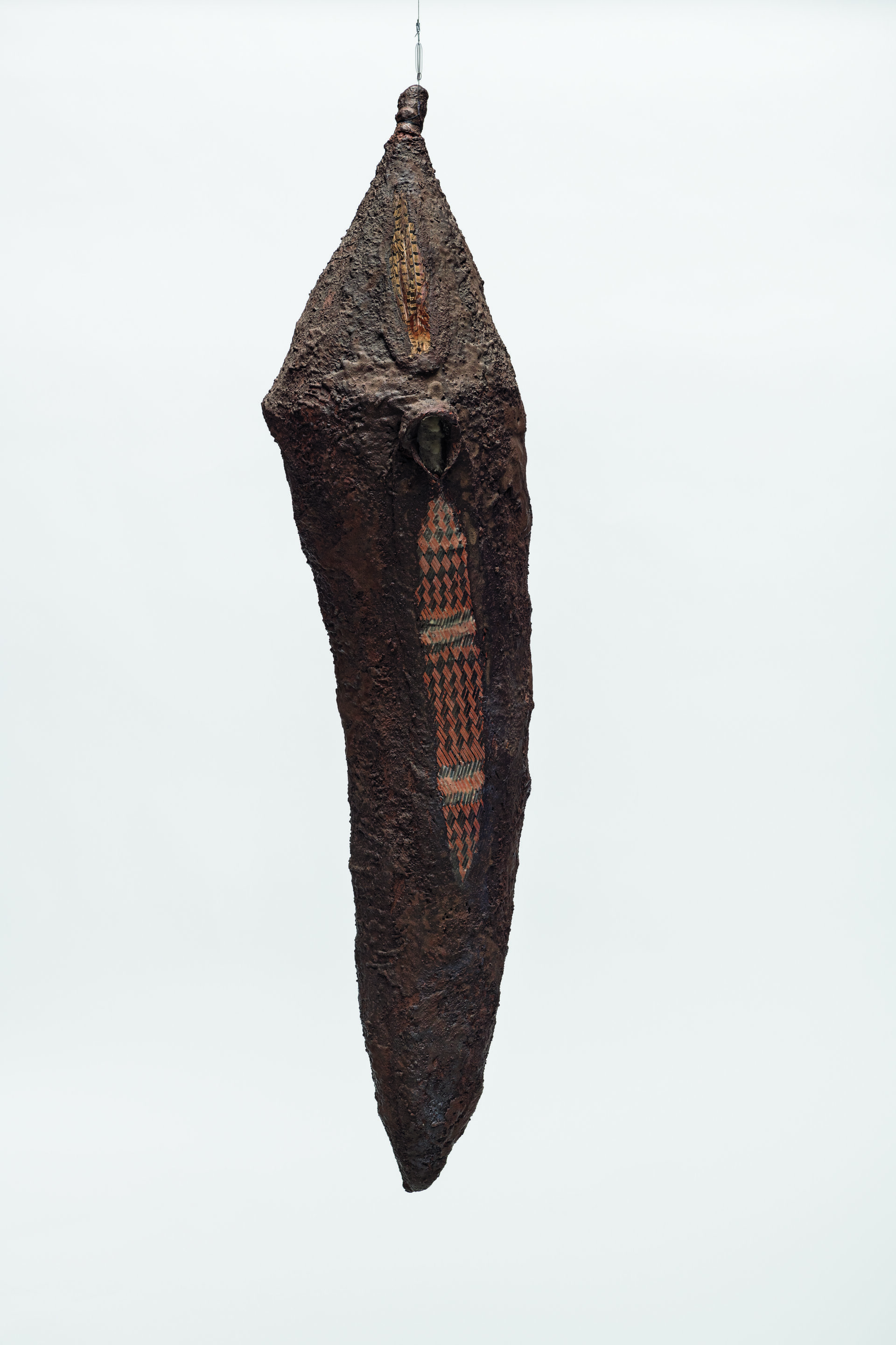 Descrição da imagem: Escultura em forma de casulo, posicionada na vertical e suspensa por um fio. A peça apresenta contornos assimétricos e alongados, com a parte superior mais larga e pontiaguda e a base afilada. A superfície é áspera e irregular, na cor marrom. No centro do corpo, uma pena de faisão, com pequenos losangos alaranjados e pretos dispostos ao longo do comprimento. Acima dela, um volume arredondado se projeta levemente para fora. Fim da descrição.
