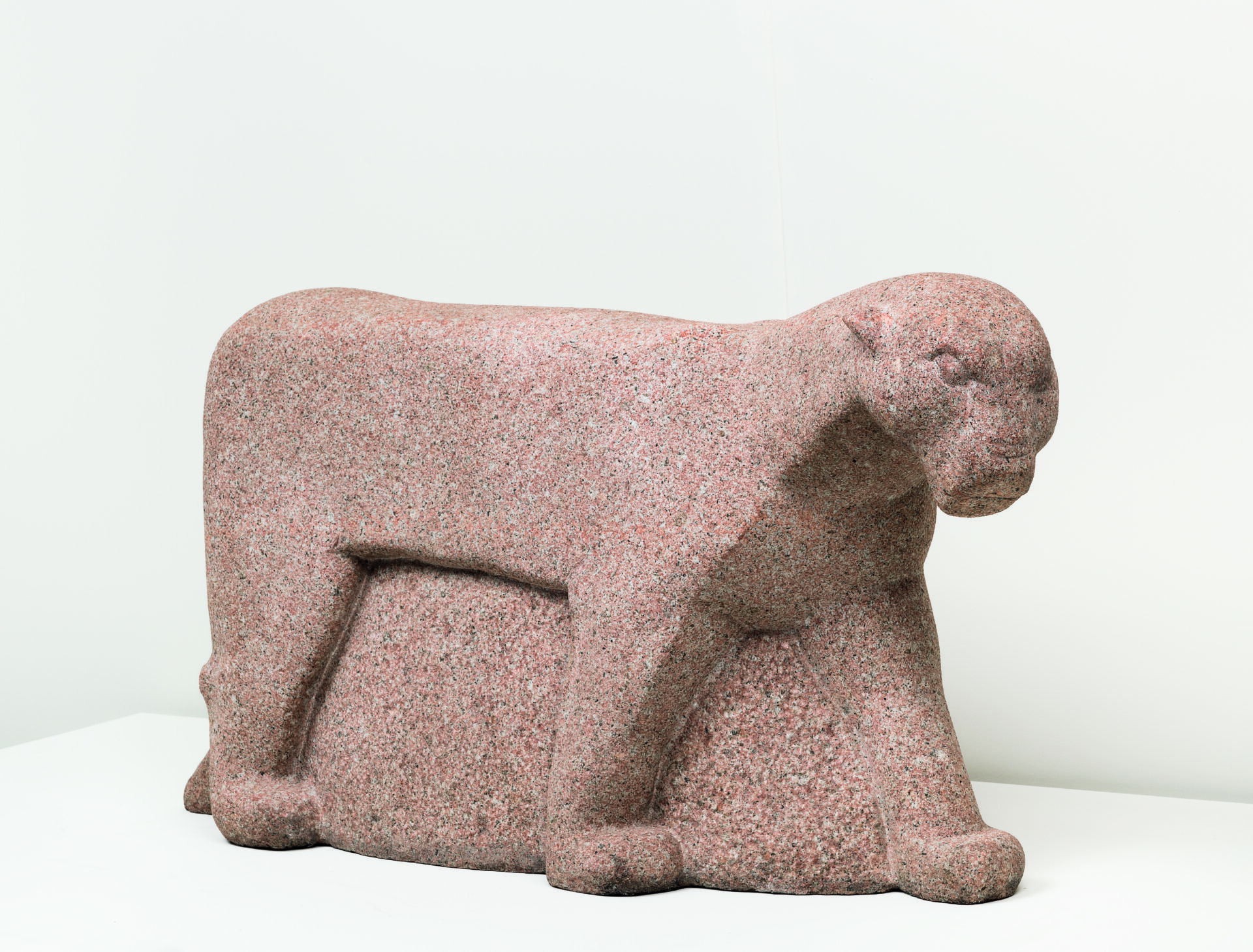 Descrição da imagem: Escultura de uma onça esculpida em granito, apoiada nas quatro patas. O corpo do animal é alongado, com formas simplificadas e geométricas, marcadas por linhas suaves e contornos arredondados. A cabeça é arredondada, com o focinho achatado. As patas são robustas e de formato retangular. A superfície apresenta textura granulada nas cores bege, branca e preta. Fim da descrição.
