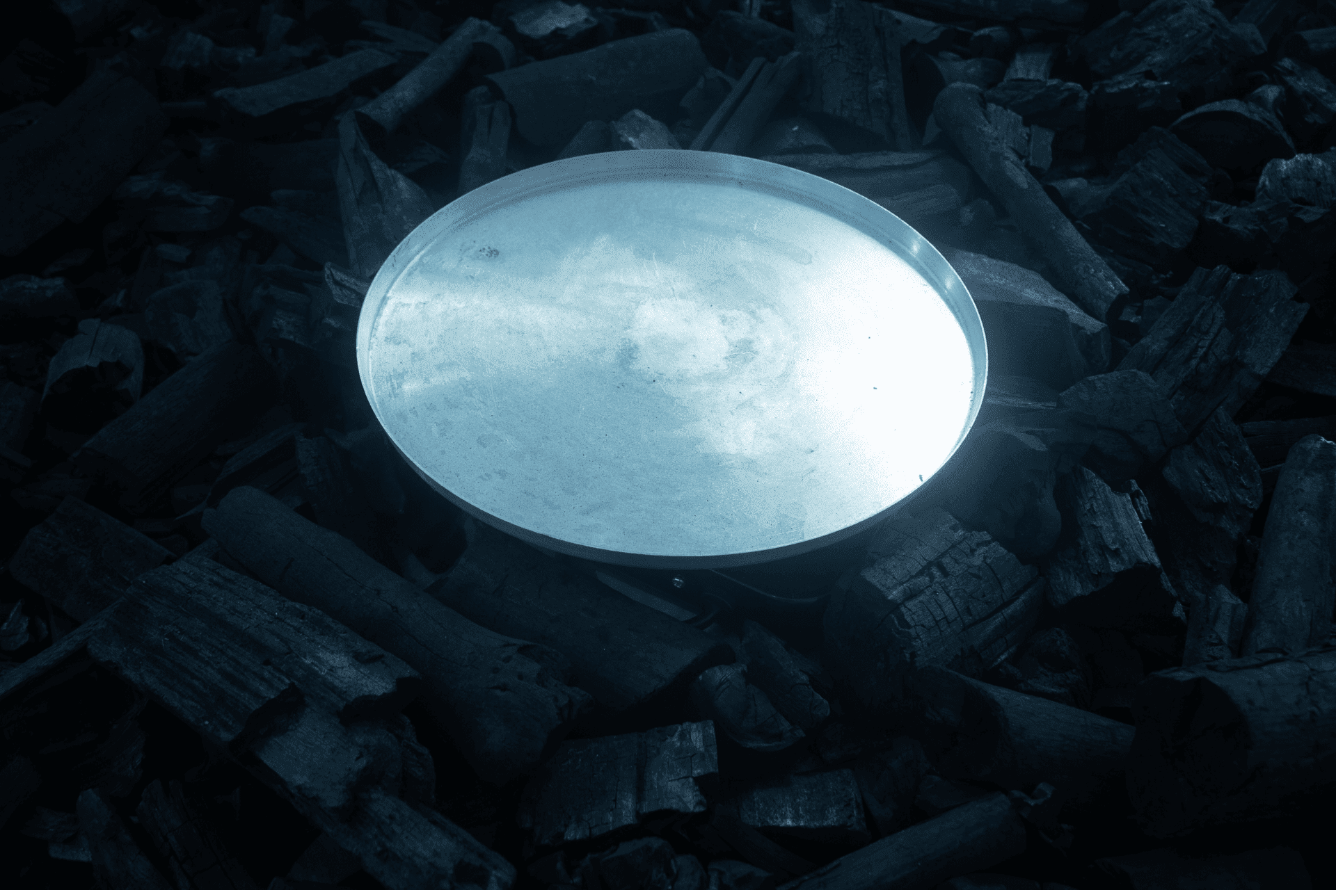 Descrição da imagem: Fotografia horizontal de parte da obra “Acender o Silêncio”. Ao centro sobre amontoado de pedaços de carvão, uma peça metálica circular. Nela incide uma luz. Fim da descrição da imagem.