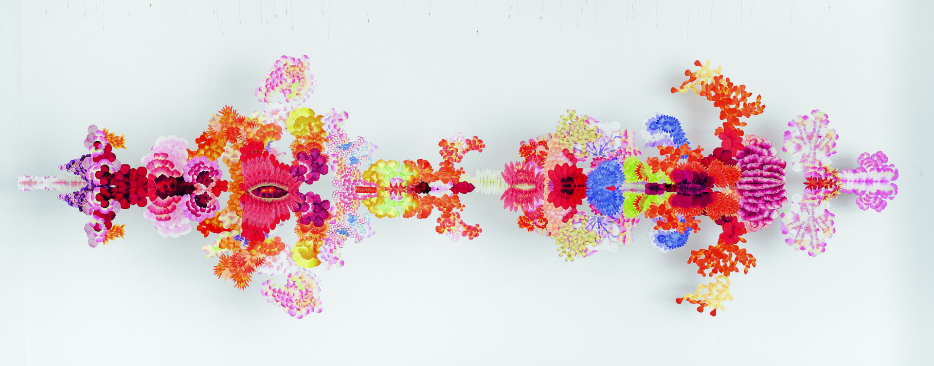 Descrição da imagem: Instalação composta por pétalas de flores artificiais coladas sobre acrílico e suspensas por fios transparentes. As flores variam em tamanho, formato e cor. Os recortes estão organizados em fileiras paralelas, intercaladas e separadas por espaços. Alguns blocos de flores se sobrepõem. Elas apresentam tons de amarelo, branco, vermelho, laranja, roxo, verde e rosa. Os formatos variam entre pétalas arredondadas, finas e pontudas. De frente, a instalação se mostra como um corpo único; de lado, revela os espaços entre os blocos que a estruturam. Fim da descrição.
