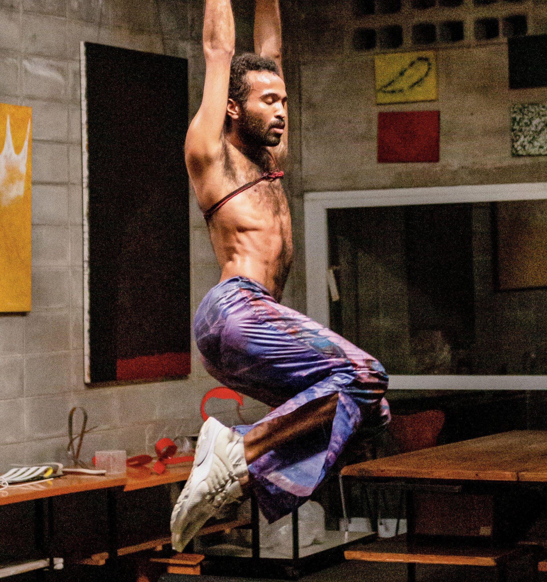 Descrição da imagem: Em outra cena da performance. Um homem negro de cabelo crespo castanho escuro e curto. Tem barba, pelos nas axilas e peito. Está suspenso pelos braços com os olhos fechados e um cordão amarrado ao peito desnudo e pernas flexionadas para trás. Usa calça estampada de azul e roxo e tênis brancos. Ao fundo, em uma parede de concreto, pequenos quadros coloridos dispostos de forma assimétrica. Fim da descrição da imagem.