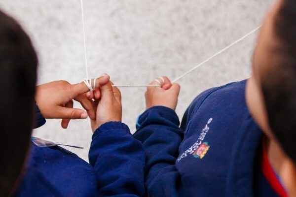 Descrição da imagem: Uma foto tirada de cima (plano plongée), focando nas mãos e braços de duas crianças vestindo uniformes azuis escuros. As crianças estão manipulando um pedaço de barbante branco entre si, criando uma figura geométrica ou uma 