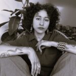 Descrição da imagem: A imagem é um retrato em preto e branco de uma mulher jovem, sentada e olhando diretamente para a câmera. Ela possui cabelos cacheados e volumosos, que emolduram seu rosto. Seus lábios são escuros e seus olhos estão fixos. A mulher veste uma camisa de manga curta com botões na frente e calças largas. Seus braços estão cruzados e apoiados sobre as pernas dobradas. No braço esquerdo, há tatuagens visíveis, incluindo uma serpente e linhas abstratas. No pulso esquerdo, ela usa uma pulseira larga com um padrão geométrico em ziguezague. No dedo anelar da mão direita, há um anel discreto. Ela também usa brincos pendurados longos e escuros. No fundo, atrás dela, há uma grande planta com folhas largas e escuras (uma figueira, possivelmente) no lado esquerdo. No lado direito do fundo, objetos desfocados e estantes são vagamente visíveis. A iluminação é suave, destacando os tons de cinza e as texturas. Fim da descrição.