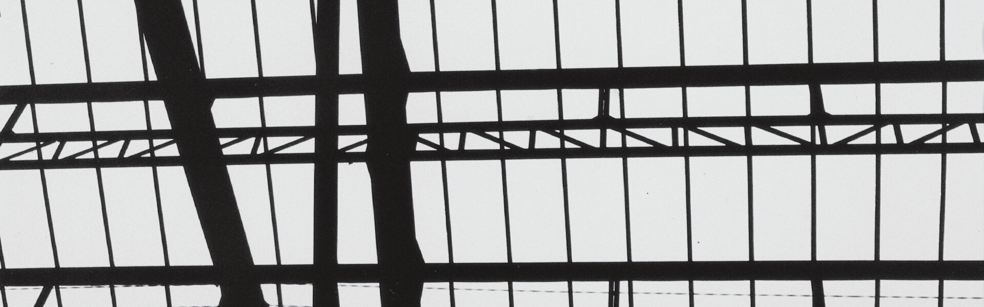 A imagem é uma fotografia em preto e branco com um estilo abstrato e geométrico, parte da série "Fotoformas" de Geraldo de Barros. Ela apresenta uma composição dominada por linhas escuras e claras, criando um padrão intrincado que lembra uma estrutura arquitetônica ou uma grade. A maior parte da imagem é preenchida por um fundo branco brilhante, que sugere um céu ou uma área de iluminação intensa. Sobre este fundo, há uma complexa rede de linhas pretas, que representam a estrutura. As linhas pretas se organizam em várias direções: Linhas Horizontais: Existem várias linhas horizontais paralelas, criando faixas ou níveis na composição. Algumas dessas linhas parecem mais grossas e contínuas, enquanto outras são mais finas. Linhas Verticais: Inúmeras linhas verticais finas cortam as horizontais, formando uma grade de retângulos ou quadrados, como se fosse uma janela, um teto de vidro ou uma ponte vista de baixo. Formas Diagonais e Irregulares: No canto inferior esquerdo e se estendendo para o centro da imagem, há um conjunto de formas pretas mais grossas e irregulares, com contornos orgânicos e curvos. Elas se cruzam e se conectam, criando a impressão de suportes, vigas ou elementos estruturais mais robustos que se destacam da grade mais fina. Estas formas maiores têm uma qualidade quase escultural, contrastando com a rigidez das linhas retas. A composição brinca com o espaço positivo (as formas pretas) e o espaço negativo (o fundo branco), e a imagem transmite uma sensação de profundidade e perspectiva, como se estivéssemos olhando através de uma estrutura complexa contra uma luz intensa. É uma fotografia que explora a geometria e a abstração através da arquitetura.