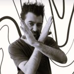 Descrição da imagem: A imagem é um retrato em preto e branco de um homem, visto da altura do peito para cima, olhando diretamente para a câmera. O homem tem cabelo escuro espetado e barba curta. Ele veste uma camiseta escura de gola redonda. O elemento central da composição são seus dois braços levantados e cruzados em frente ao rosto e peito, formando um grande "X". Suas mãos estão abertas e viradas para fora. O fundo da imagem é branco e apresenta um padrão gráfico abstrato, com linhas pretas grossas e fluidas, que se curvam e se sobrepõem. Fim da descrição.