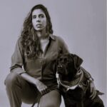 Descrição da imagem: A imagem é um retrato em preto e branco de uma mulher e um cachorro, ambos olhando para a câmera. A mulher está agachada no lado esquerdo da imagem, com o corpo ligeiramente virado para a direita. Ela possui cabelos longos e ondulados, que caem sobre seus ombros. Veste uma camisa de manga comprida com botões na frente e calças escuras. Um cinto escuro com detalhes em relevo está visível na altura de sua cintura. Sua mão direita está apoiada na perna e ela segura uma coleira escura na mão esquerda. Ao lado direito da mulher, sentado ereto, está um cachorro Labrador preto. O cachorro usa uma bandana clara com um padrão estampado em volta do pescoço e um arreio escuro. O braço esquerdo da mulher está suavemente repousado sobre o corpo do cachorro. O fundo da imagem é uma parede lisa de cor clara e uniforme. A iluminação é suave, destacando os contornos e texturas do cabelo da mulher, do pelo do cachorro e das roupas. Fim da descrição.