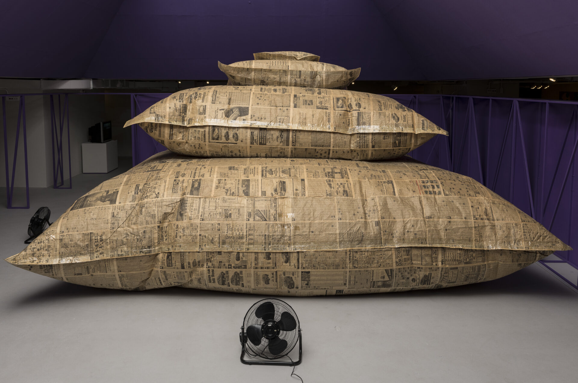 Descrição da imagem: Escultura formada por quatro almofadas infladas em grande escala, empilhadas em formato de pirâmide. As de baixo são maiores que as de cima. Todas são feitas de folhas de jornal amareladas, com anúncios, reportagens e propagandas impressos em preto. Há dois ventiladores de chão pretos, um em cada uma das laterais da obra. Está sobre um chão cinza. Fim da descrição. 
