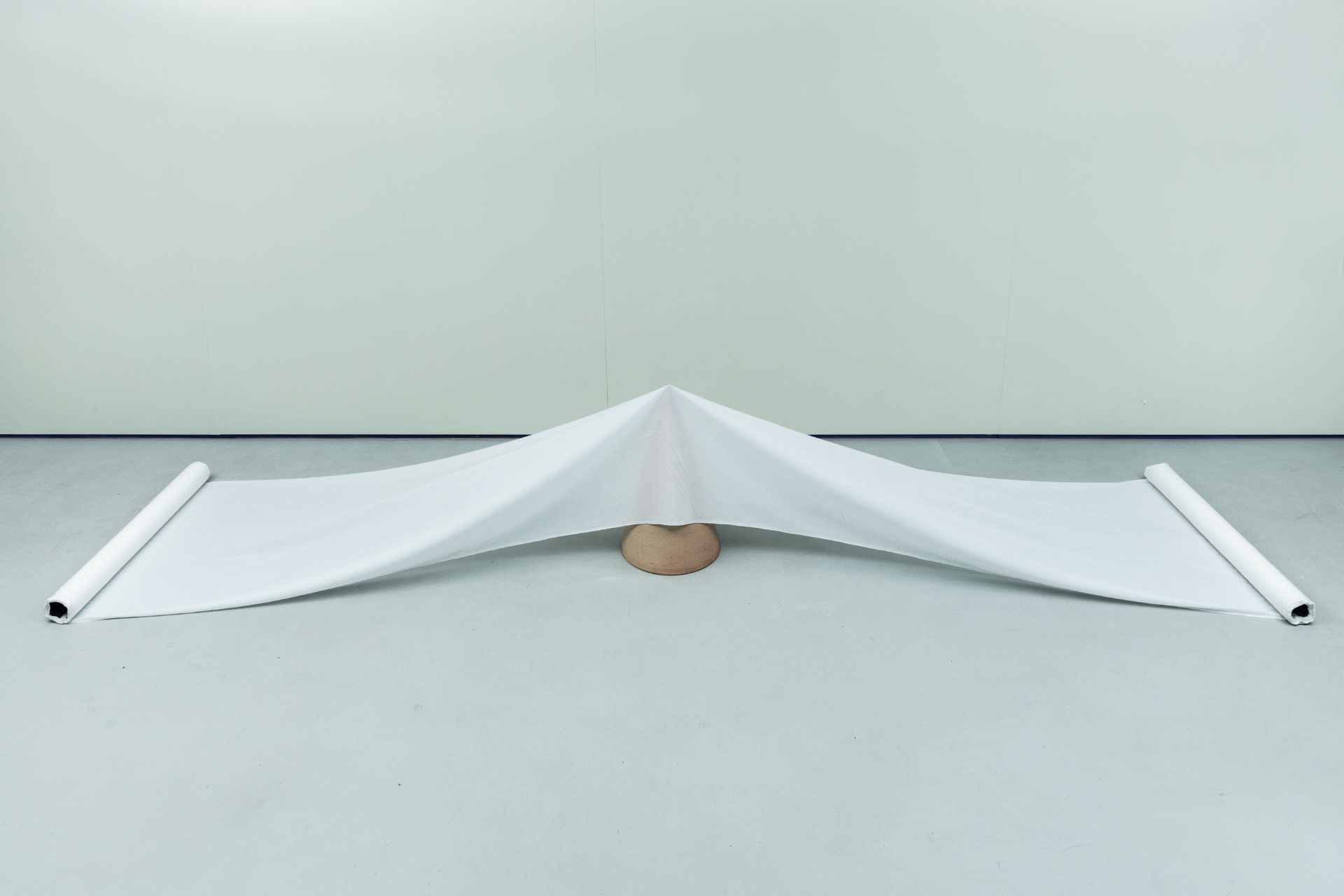 Descrição da imagem: Escultura em cerâmica, ferro e tecido. No centro, no chão, um pequeno cone de cerâmica com a ponta para cima sustenta um tecido branco translúcido, que o cobre parcialmente e se estende pelo chão para ambas as laterais. Nas extremidades, o tecido está enrolado em tubos metálicos. Fim da descrição. 
