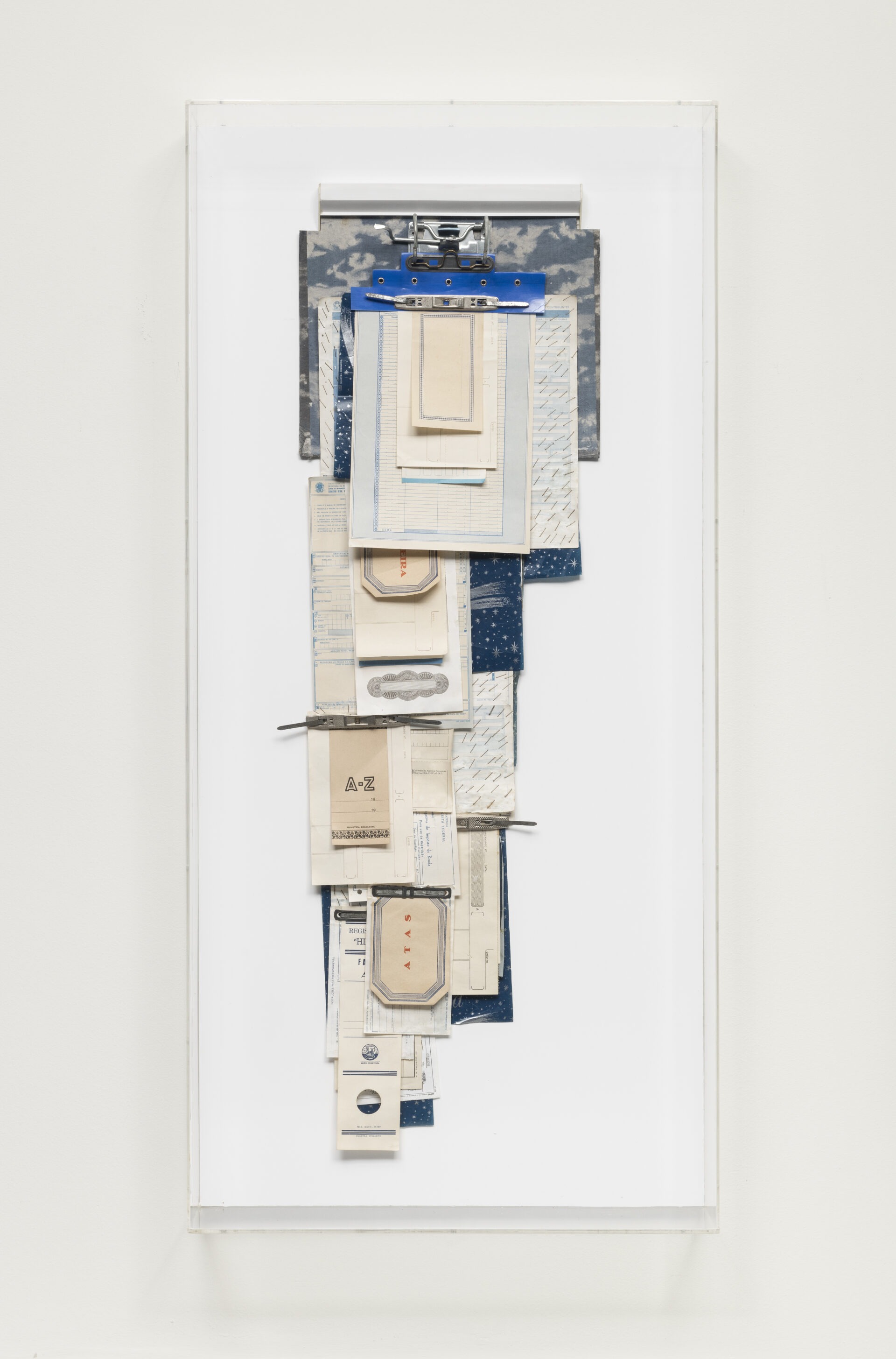 Descrição da imagem: Assemblage de formato vertical irregular, fixada em fichário. Obra criada a partir da combinação de objetos diversos. É composta por papéis, grampos trilho metalizados e papel carbono, sobrepostos e organizados em quatro camadas e tamanhos diferentes.
Na parte superior, fixado em uma moldura branca com fundo branco, o mecanismo metálico de um fichário prende uma base de papel azul, sobre a qual um grampo trilho fixa diversos papéis sobrepostos em bege e azul. Algumas dessas peças têm linhas impressas, padrões geométricos e pequenas inscrições. De cima para baixo, os papéis vão dos mais largos aos mais estreitos. Fim da descrição. 
