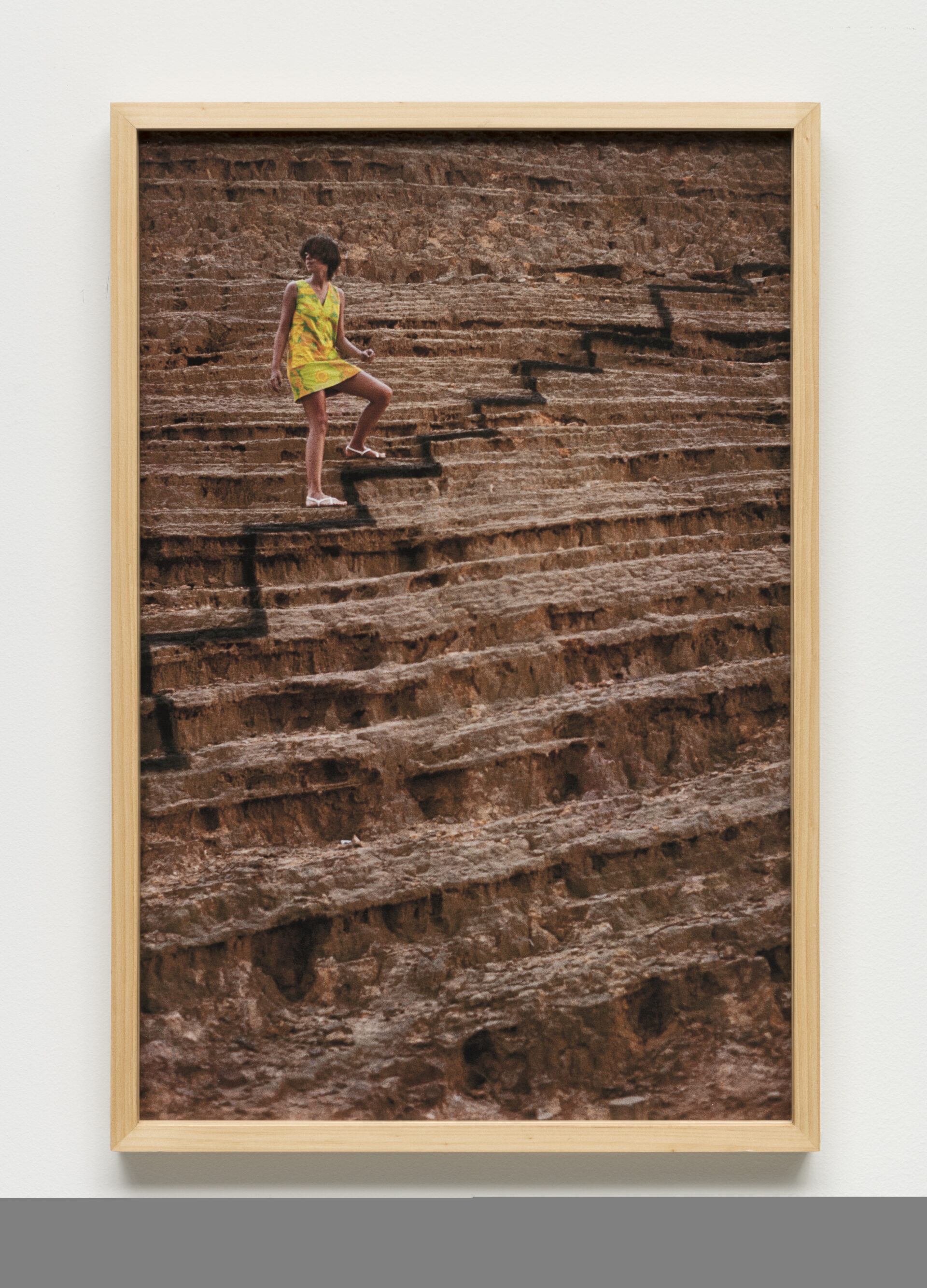 Descrição da imagem: Foto de uma mulher sobre degraus de pedra em ruínas. A mulher é parda, tem cabelos pretos, curtos e lisos, com franja que cobre parcialmente os olhos. Ela usa um vestido verde, curto, de alça larga, acima dos joelhos e sandálias brancas. Está com o rosto virado para a esquerda e o corpo para frente. Um pé está sobre um degrau e o outro, sobre o degrau acima. Há um risco preto sobre os degraus de pedra à frente dela, formando o desenho de outros degraus. A moldura é bege. Fim da descrição. 
