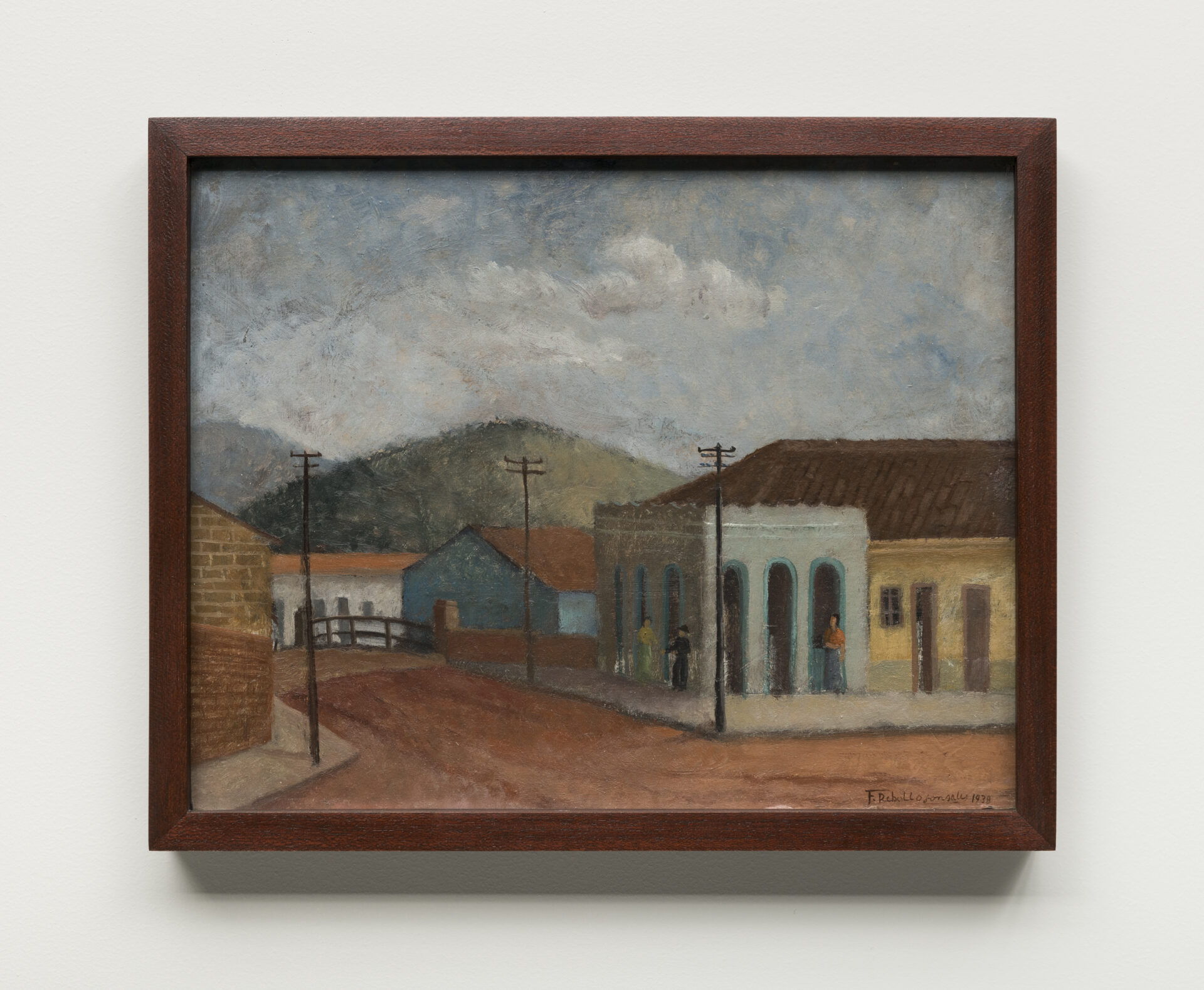 Descrição da imagem: Pintura de uma esquina com casas nas cores amarela, branca e azul, todas com telhados marrons. Algumas portas são retangulares, outras em arco. Três postes de energia estão distribuídos pelas calçadas.
À direita, sobre a calçada de uma das casas, pintada de azul, três pessoas de pele bege representadas apenas pelo contorno do corpo: duas frente a frente e uma mais afastada. A primeira usa camiseta amarela e saia verde; a segunda, chapéu, camisa e calça pretos; a terceira, camiseta laranja e saia azul. A rua é de terra batida avermelhada. Ao fundo, uma colina esverdeada sob céu claro com nuvens brancas. No canto inferior direito, há uma pequena assinatura em preto: F Rebolo Gonsales. Mil novecentos e trinta e oito. A moldura é marrom. Fim da descrição. 
