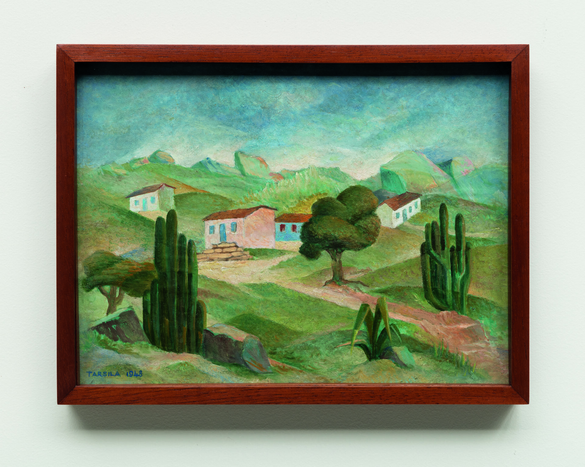 Descrição da imagem: Pintura de uma paisagem rural em tons suaves de verde, azul e marrom. O terreno é coberto por grama verde. À frente, na parte inferior de cada lateral, há um conjunto de cactos verticais e finos em tons de verde-escuro. Uma estrada sinuosa de terra batida corta o terreno do canto inferior direito até o centro da pintura, onde está uma árvore de copa arredondada. Atrás da árvore, sobre pequenas elevações irregulares, há quatro casas: uma branca, uma marrom-clara, uma azul e outra branca, todas com telhados marrons. Ao fundo, há montanhas em formato arredondado, sob um céu azul esfumado. No canto inferior esquerdo, há uma pequena assinatura em azul-turquesa: Tarsila mil novecentos e quarenta e oito. A moldura é marrom. Fim da descrição. 
